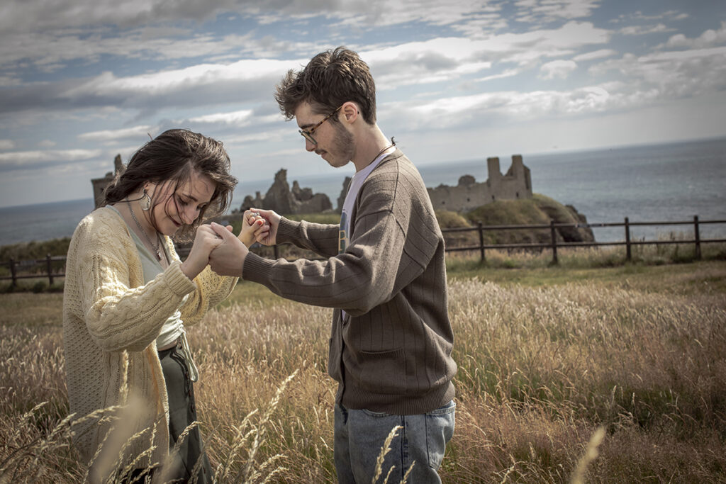 A transgender couple having an engagement session in-front of Dunnotter Castle in Scotland. The couple are wearing woollen sweaters, dancing and laughing in the wind.
