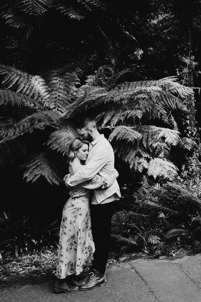 Black and white photo of a couple embracing in the gardens in Mt Dandenong. The couple are having an engagement session in order for them to feel more relaxed on their wedding day. He is kissing her forehead.