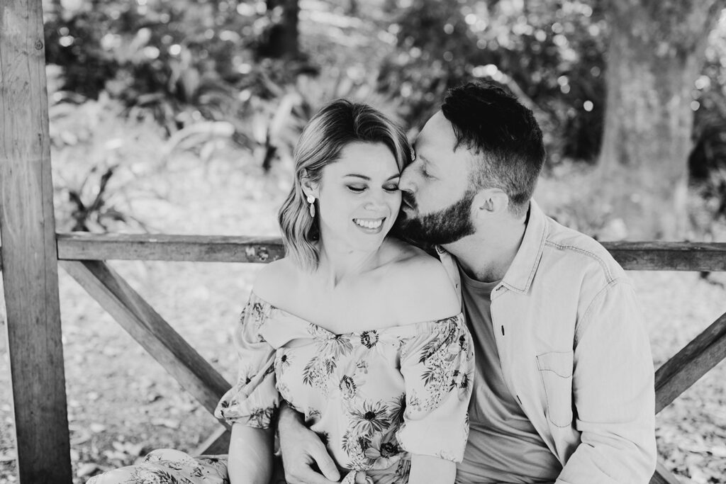 groom kissing bride on the cheek as they sit side by side in a park, feeling relaxed and more confident. 
Black and white photo of a couple having an engagement session in Mt Dandenong.
She's wearing an off the shoulder floral dress.