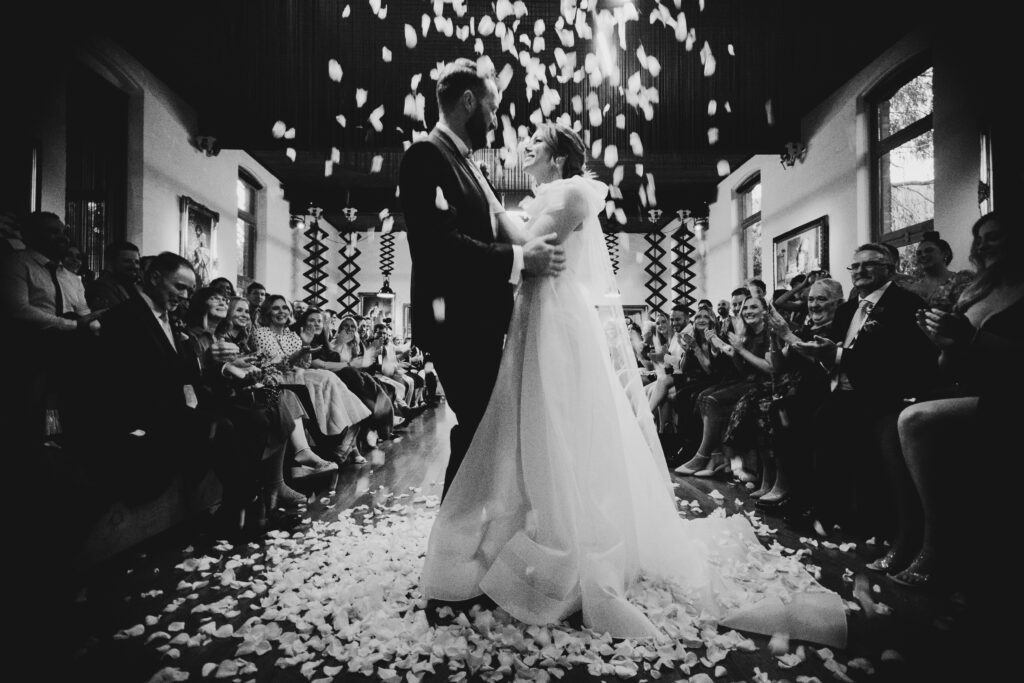 bride and groom in a black and white image standing under a shower of rose petals during first kiss of ceremony