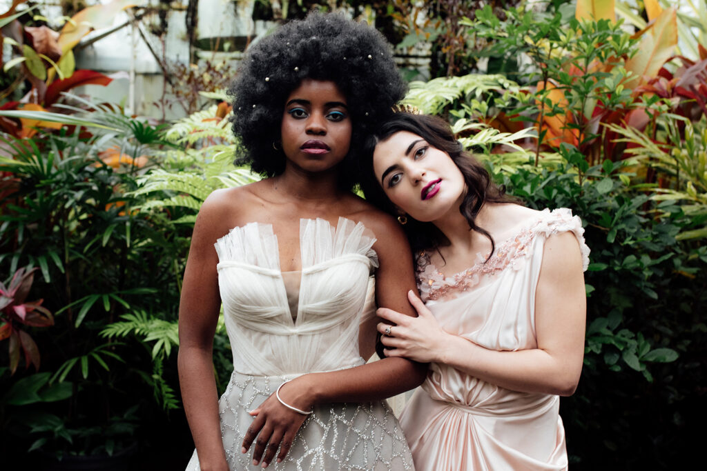 Colour image of 2 girls standing in a greenhouse wearing gorgeous gowns after having their makeup done by one of the best wedding makeup artists in Melbourne. Photograph taken by Liminal wonderland wedding Photographer
