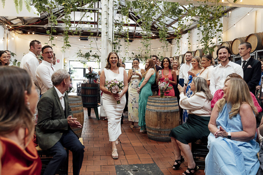 A Bride waling down the aisle at Noisy Ritual urban winery. She is glowing and happy and smiling at all her friends.