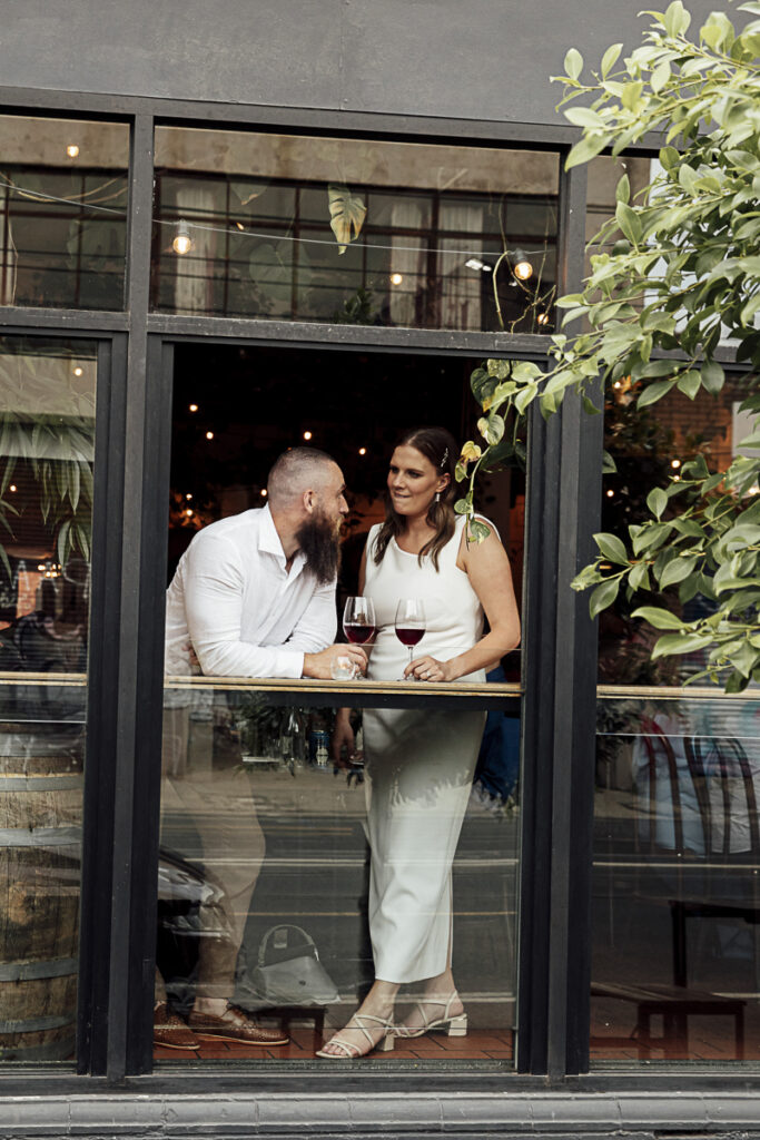 A bride and groom standing at a window sharing a glass of wine at Noise Ritual