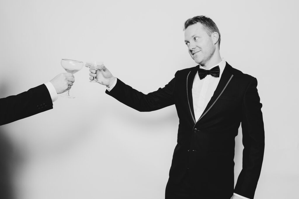 Black and white photo of a man standing in a tuxedo clinking his glass ina. cheers have a photo taked