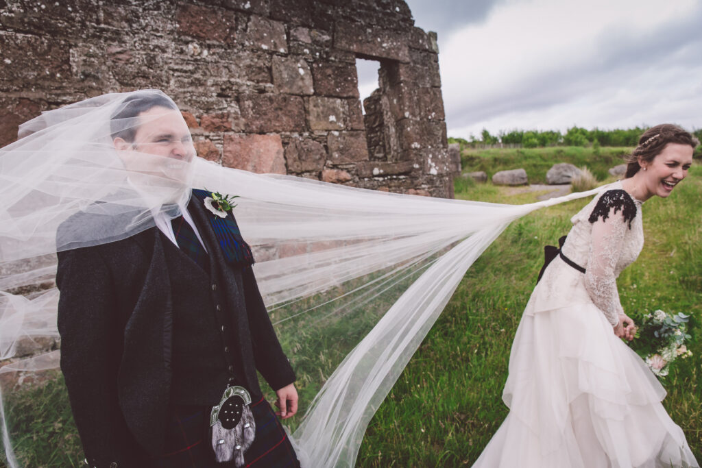 a couple on their wedding day having photos taken in Scotland. She's wearing a custom wedding dress with tulle and a cape veil very flowy