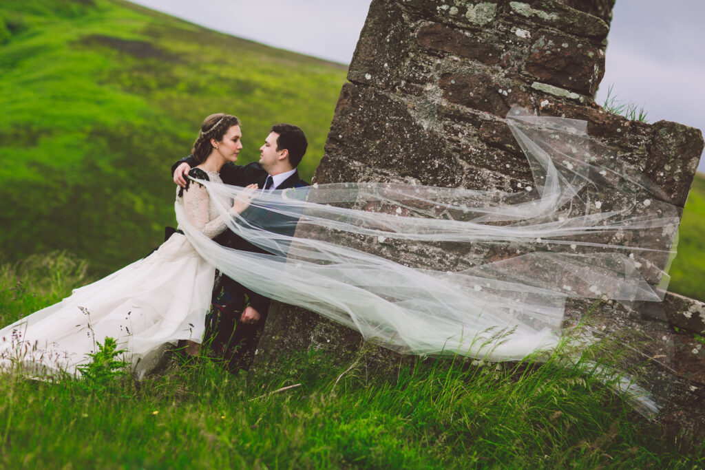 a couple on their wedding day having photos taken in Scotland. She's wearing a custom wedding dress with tulle and a cape veil very flowy