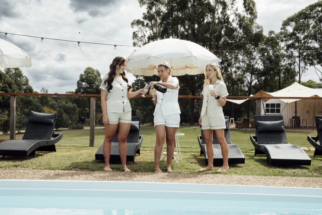 A colour image of a bride with her bridesmaids, stanifn by the swimming pool at eight acres in lakes entrance. They are popping a bottle of Champagne and laughing.