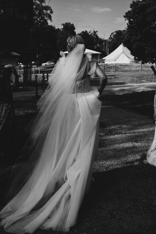 A black and white image of a bride walking in her wedding dress from the back at Eight Acres Lakes Entrace