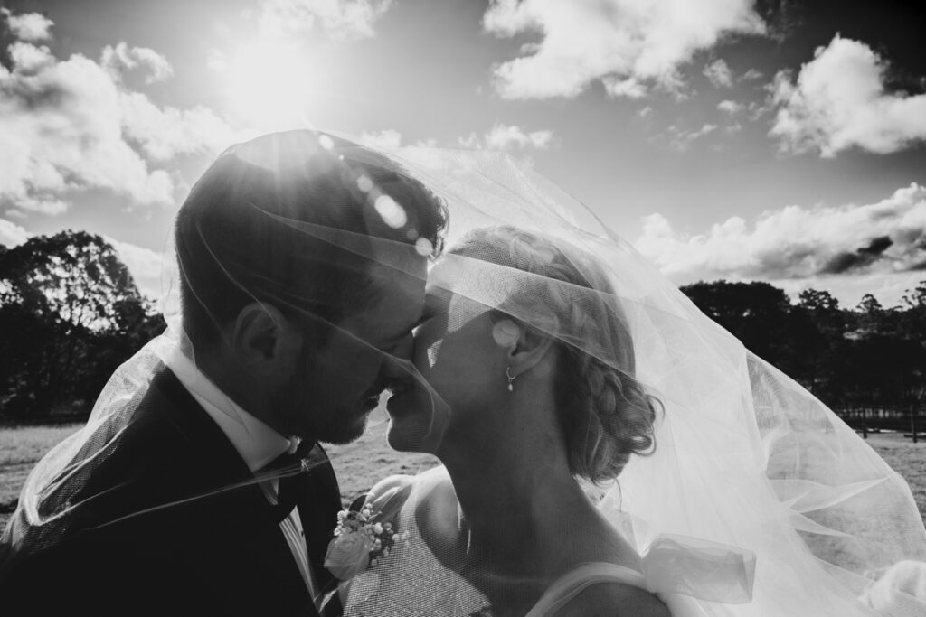 A black and white image of a couple kissing with a veil wrapped around them at lakes Entrance. Eight Acres