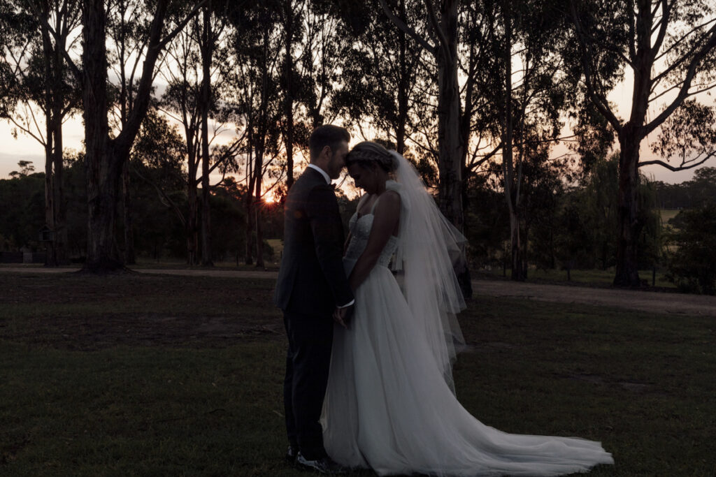 a colour image of a bride and groom at sunset, the gum trees are sihoetted in the background. They are at Eight Acres Lakes Entrance.