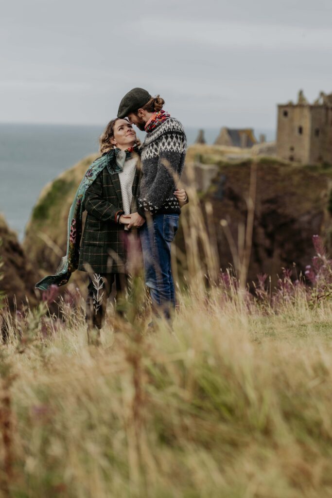 A couple having wedding photos taken near Aberdeen, at Dunnotter castle in Scotland. They are walking through the flowery fields. She is wearing a Helen Ruth scarf