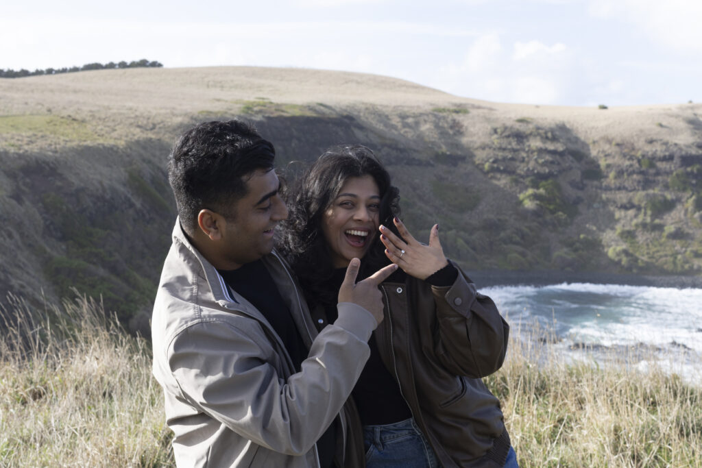 Melbourne Proposal Photography a couple getting engaged in Flinders. Young couple standing on cliff top laughing and pointing at ring