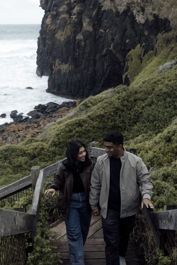 Melbourne Proposal Photography a couple getting engaged in Flinders. Young couple standing on cliff top laughing, holding hands and walking