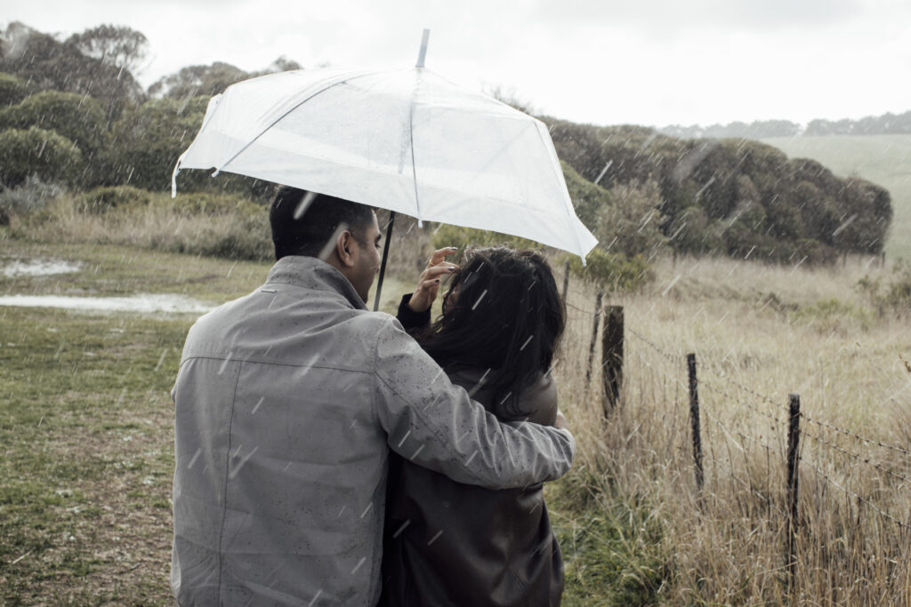 Melbourne Proposal Photography a couple getting engaged in Flinders. Young couple standing on cliff top laughing, holding hands and walking in the rain under an umbrella