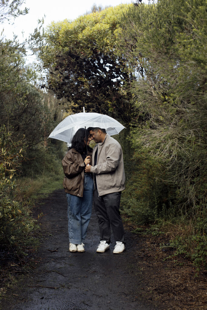 Melbourne Proposal Photography a couple getting engaged in Flinders. Young couple standing on cliff top laughing, holding hands and walking in the rain under an umbrella