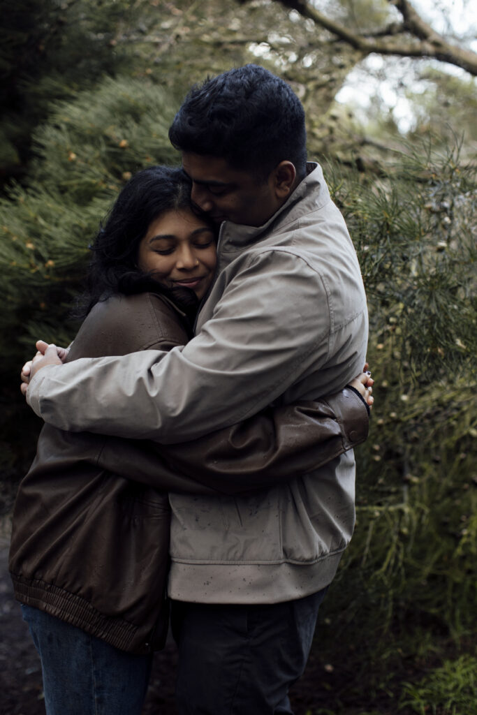 Melbourne Proposal Photography a couple getting engaged in Flinders. Young couple standing on cliff top laughing, holding hands and walking in the rain under an umbrella