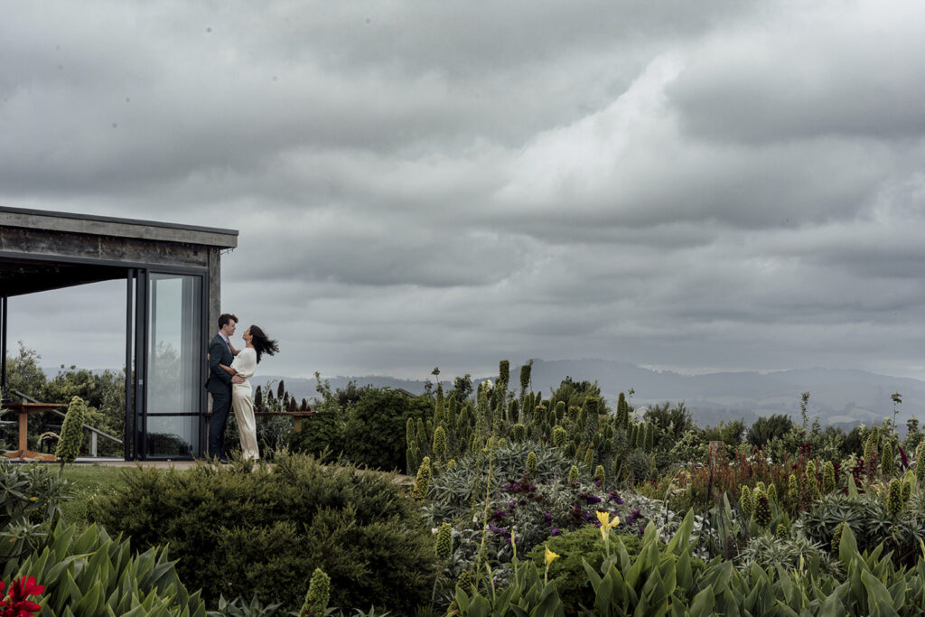 A couple having portraits at Gurney's cider. The girl wears a beautiful gold jumpsuit, windswept hair. They are snuggly and close standing the sky is dark and moody and stormy. Standing with Wilsons prom in the background.