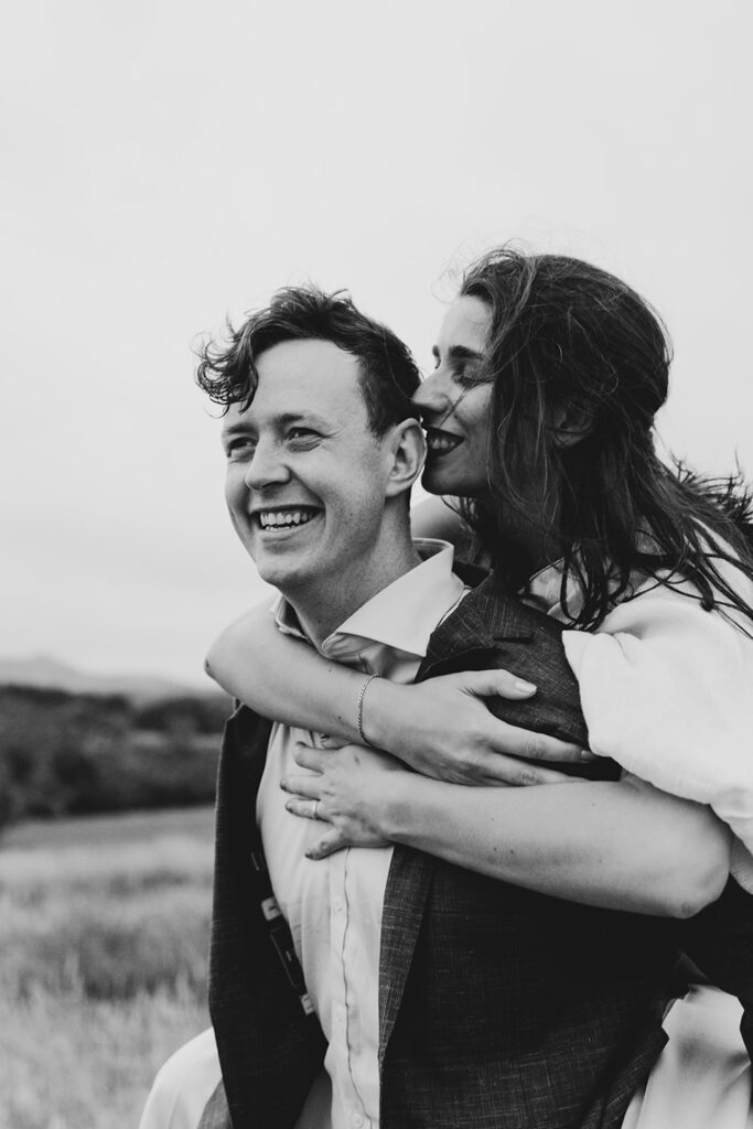A couple having portraits at Gurney's cider The girl wears a beautiful gold jumpsuit, windswept hair. They are snuggly and close standing in the long grass, the sky is dark and moody and stormy. Black and white photo laughing and having fun.
