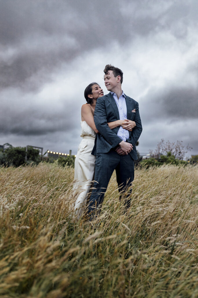 A couple having portraits at Gurney's cider. The bride wears a beautiful gold jumpsuit, windswept hair. They are snuggly and close standing in the long grass, the sky is dark and moody and stormy.