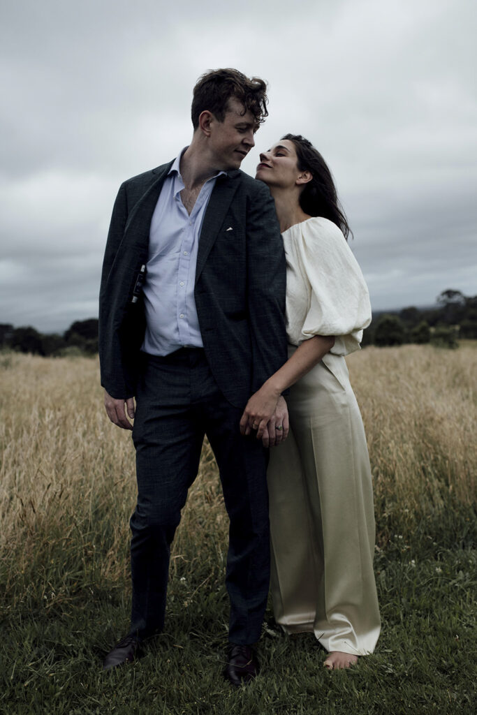 A couple having portraits at Gurney's cider. The girl wears a beautiful gold jumpsuit, windswept hair. They are snuggly and close standing in the long grass, the sky is dark and moody and stormy. The couple are holding hands looking longingnly at each other