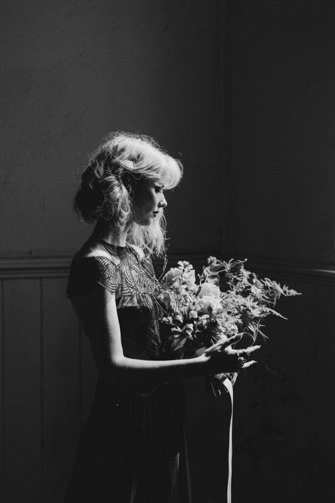A black and white photo of a bride on her wedding day, it is dramatically lit with the sun streaming through the windows, she is looking down at her bouquet of flowers.