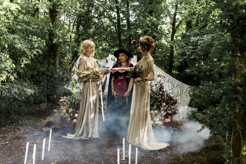 A wedding photo of two brides at Scrub Hill, they are having a spiritual led ceremony with Celebrant Jam. There is smoke and candles in the background making the image more etherial. They brides are wearing long gold silk gowns by Gwendolyn.