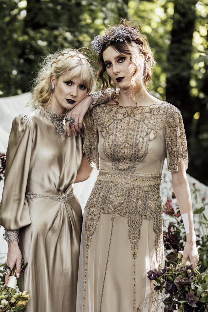 two brides on their wedding day at Scrub Hill, they are wearing silky golden dress with a 1920's Gatsby vibe from Gwendolyn couture in Melbourne. They are wearing beautiful silver crowns from Stephanie Browne. They are leaning on each on each, their expression is still and looking a tthe camera.
