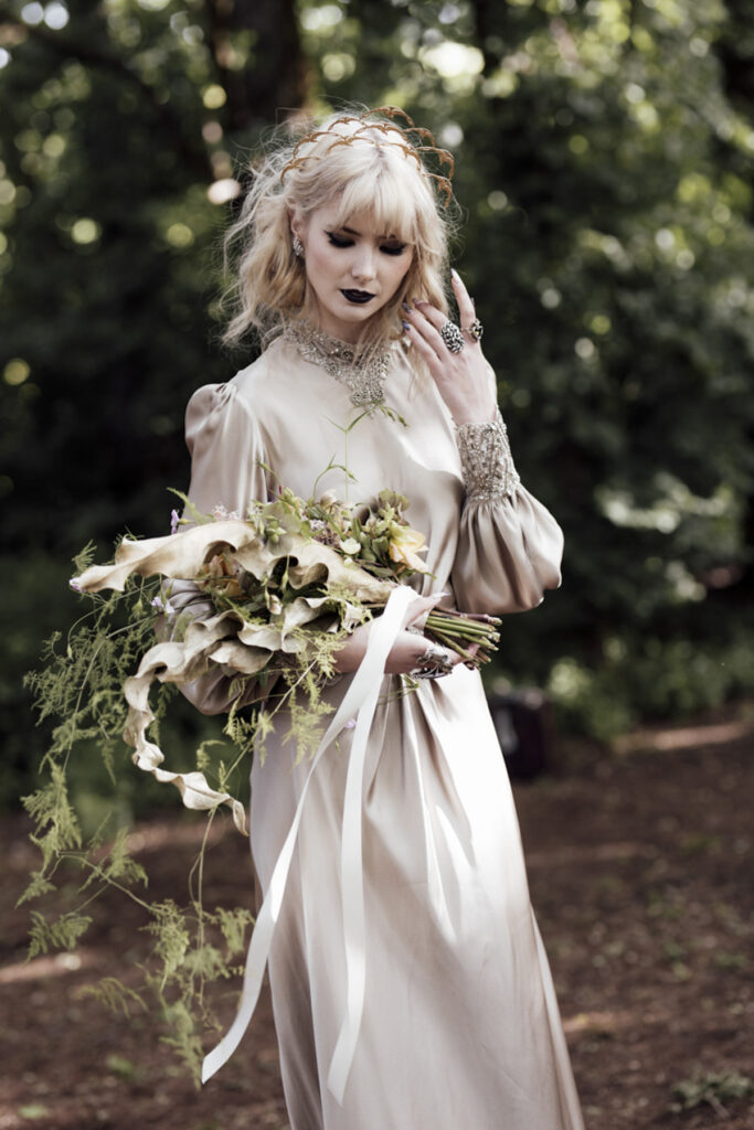 a bride walking through the forest at Scrub Hill on her wedding day, it is softly lit, alternative and etherial looking with a green hue. She is wearing a long golden silk gown with a Gatsby vibe from Gwendolyn Couture