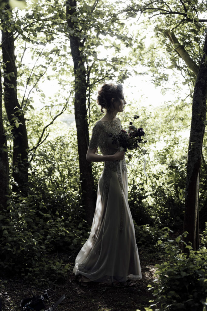 a bride walking through the forest at Scrub Hill on her wedding day, it is softly lit, alternative and etherial looking with a green hue. She is wearing a long golden silk gown with a Gatsby vibe from Gwendolyn Couture