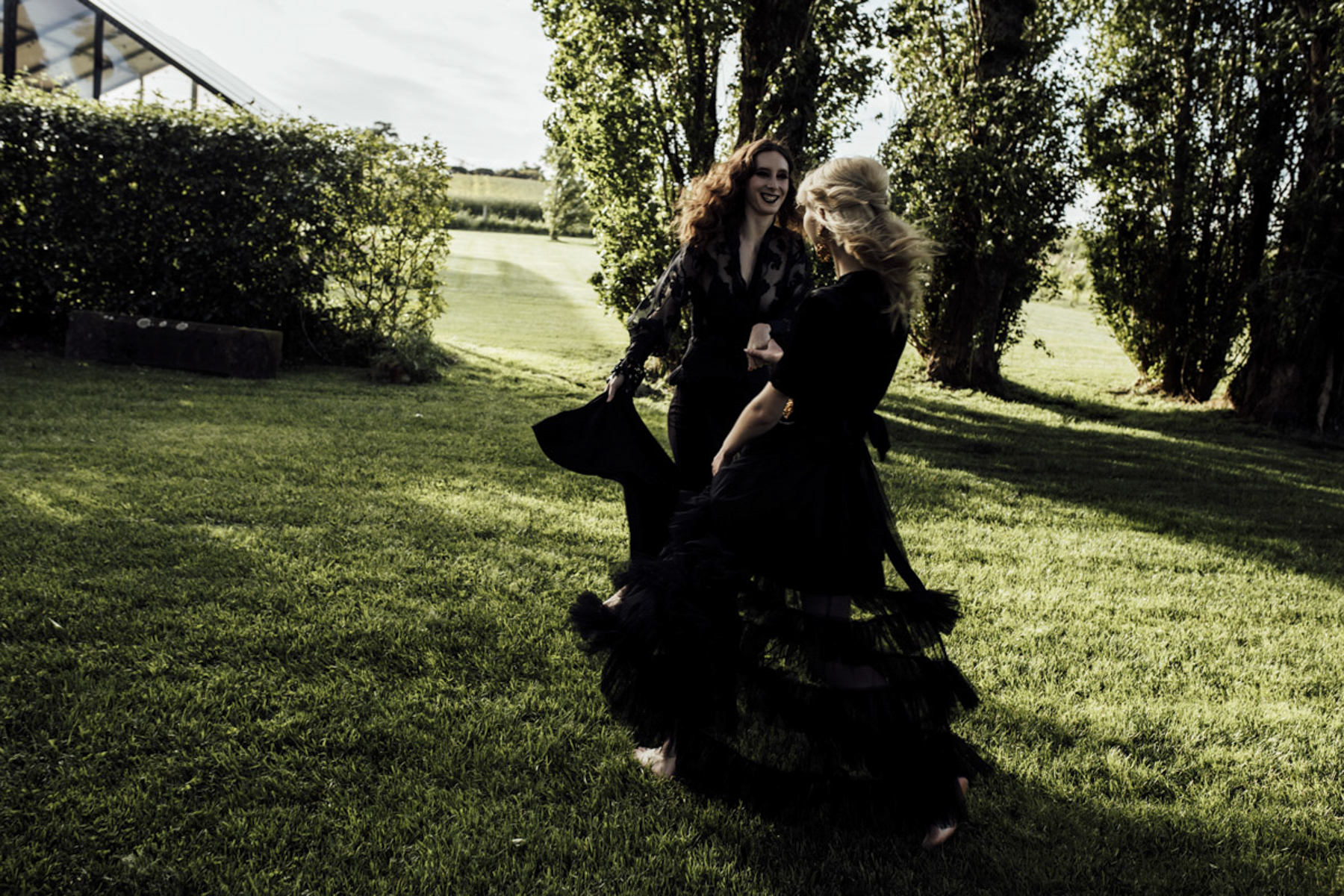 a photograph of two women dressed in black beautiful dresses, dancing at Scrub Hill on a wedding day. It's a more alternitive LGBTQI+ vibe. The green grass is lush and vibrant behind them, shot by Liminal Wonderland wedding photographer