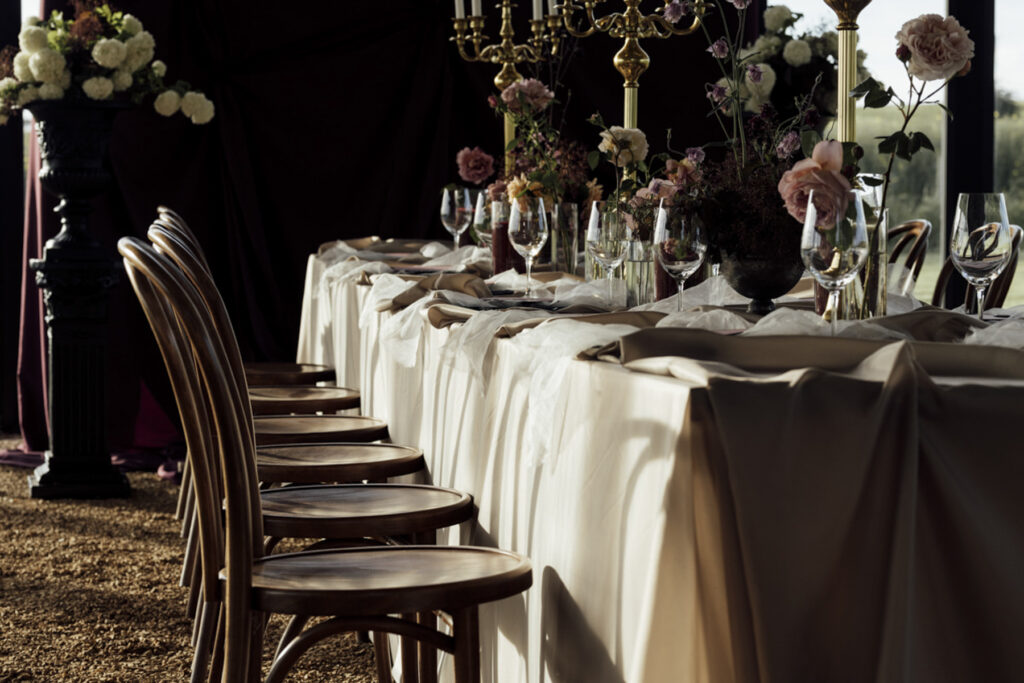 Wedding reception table setting at Scrub Hill, The table linen is silk and glowy golden, the flowers are soft and warm, candelabras line the table to give a moody vibe. The overall image is warm and lucious