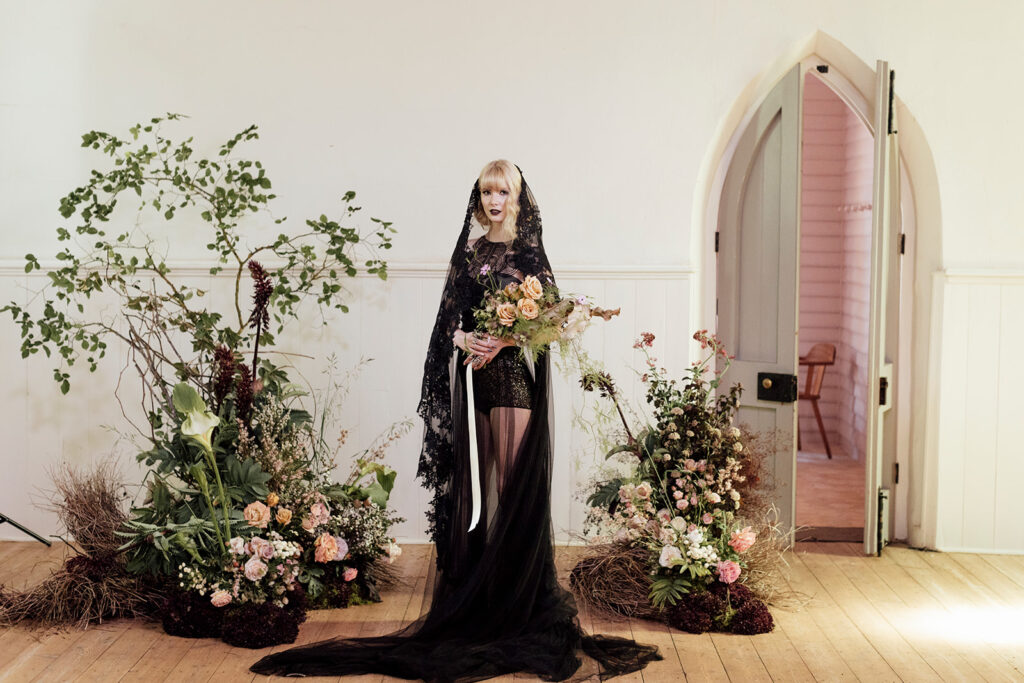 Beautiful flowers from Larkspur Flowers. The flowers are wild and beautiful and soft looking. A bride stands in the middle of all the sculptural flowers she is dressed in a black wedding dress, with a veil that looks slightly spannish in vibe.