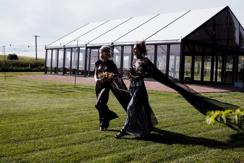 two women at a wedding in Scrub Hill, they are wearing black gowns by Gwendolyn Couture. They alos have incredible crowns by Stephanie Browne, the flowers are wild by Larkspur flowers. The setting is beautiful and summery.