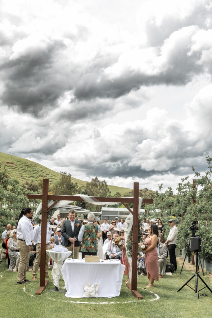 ceremony space at DV Cider, the blossoms are in full bloom, there is an arbour and a bride and groom getting married, exchanging vows in an emotional led wedding.