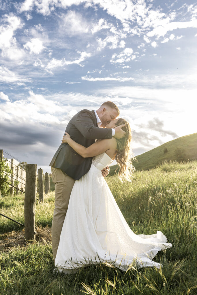 Colour image of a couple in the orchard at DV Cider. they are surrounded by trees in blossom at golden hour they are embracing one another and the groom is kissing her on the forehead. The bride hair is down and she is in a white gown