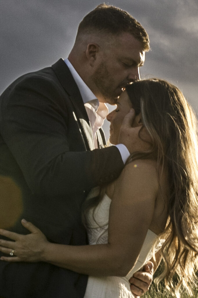Colour image of a couple in the orchard at DV Cider. they are surrounded by trees in blossom at golden hour they are embracing one another and the groom is kissing her on the forehead. The bride hair is down and she is in a white gown