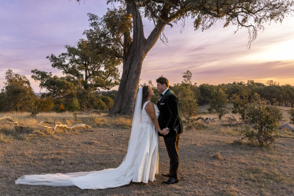 A colour wedding photo of a bride and groom sharing a moment at sunset, they are at the top of the hill at Paramoor standing close with her flowers in the middle the wind has cought her veil. The sky is lit with oranges and blue hues.