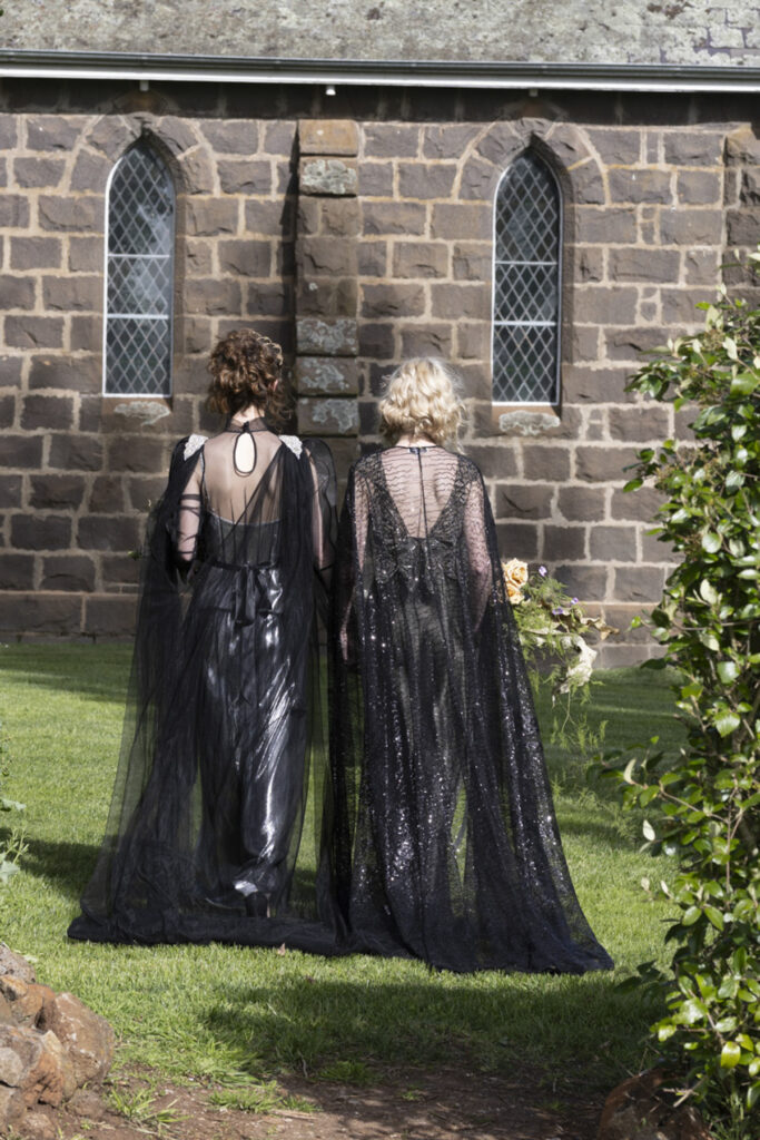 two women at a wedding in Scrub Hill, they are wearing black gowns by Gwendolyn Couture. They alos have incredible crowns by Stephanie Browne, the flowers are wild by Larkspur flowers. The setting is beautiful and summery. The photo is set against the blue stone chapel of Scrub Hill