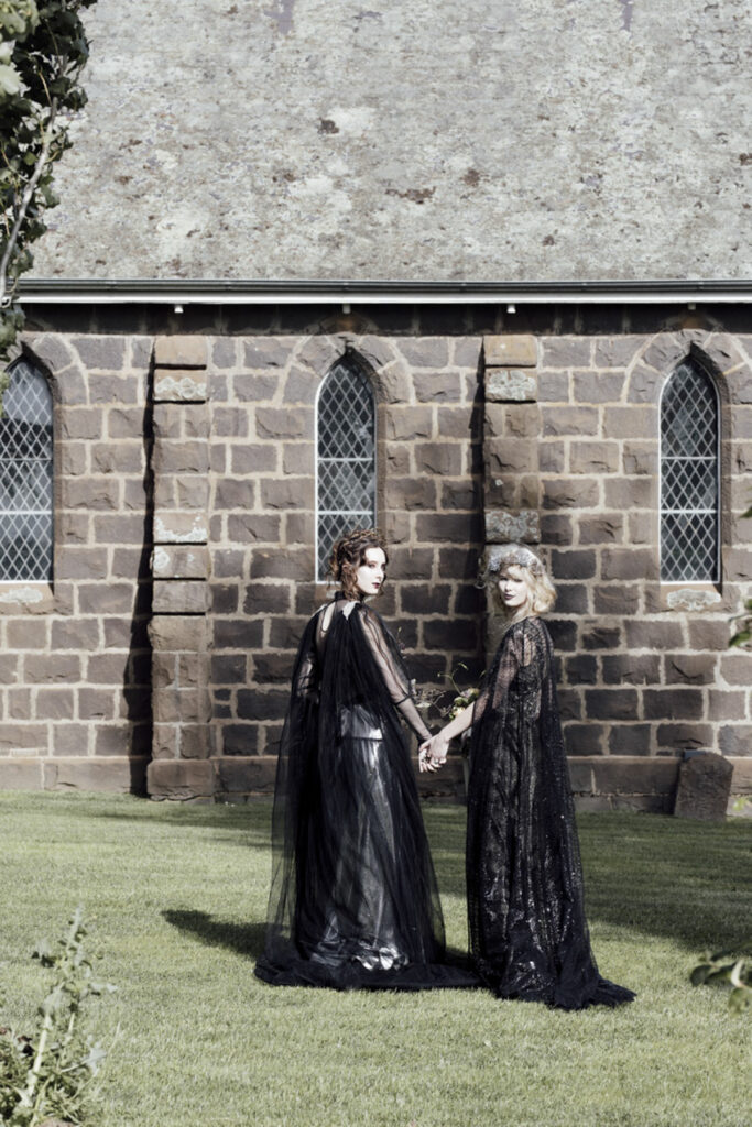 two women at a wedding in Scrub Hill, they are wearing black gowns by Gwendolyn Couture. They als0 have incredible crowns by Stephanie Browne, the flowers are wild by Larkspur flowers. The setting is beautiful and summery. The photo is set against the blue stone chapel of Scrub Hill