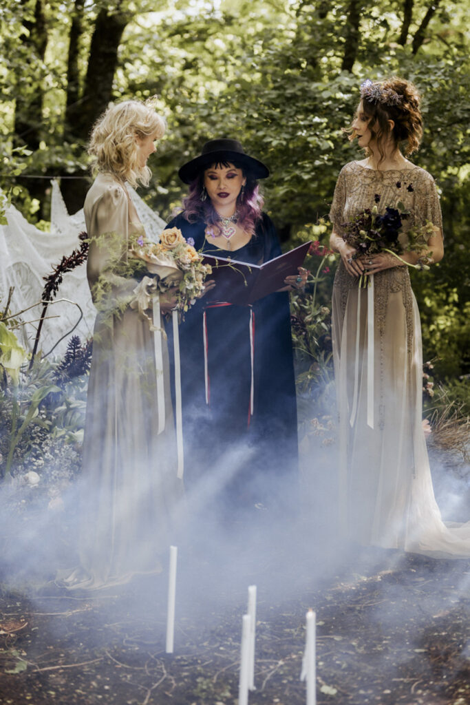 A wedding photo of two brides at Scrub Hill, they are having a spiritual led ceremony with Celebrant Jam. There is smoke and candles in the background making the image more etherial. They brides are wearing long gold silk gowns by Gwendolyn.