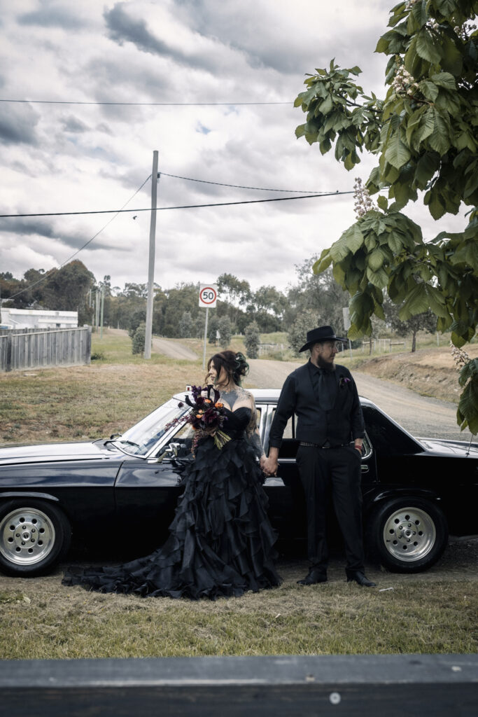 A bride and groom in black car, the bride is in a black ruffle wedding dress with dark moody flowers. The couple are more alternative with tattoos.
