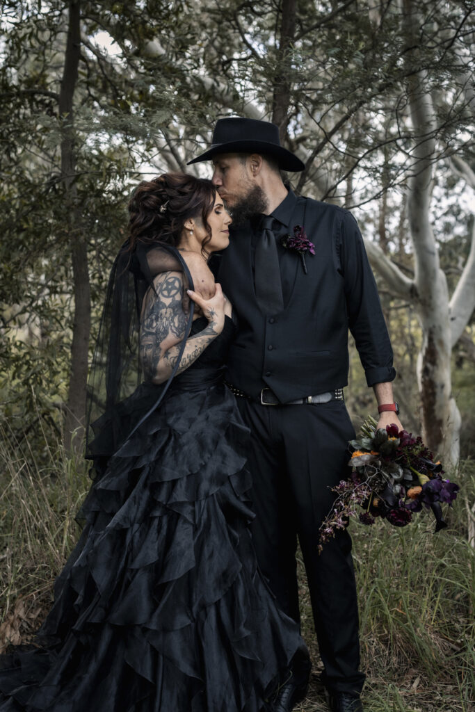 A bride and groom in black outfits, the bride is in a black ruffle wedding dress with dark moody flowers. The couple are more alternative with tattoos.