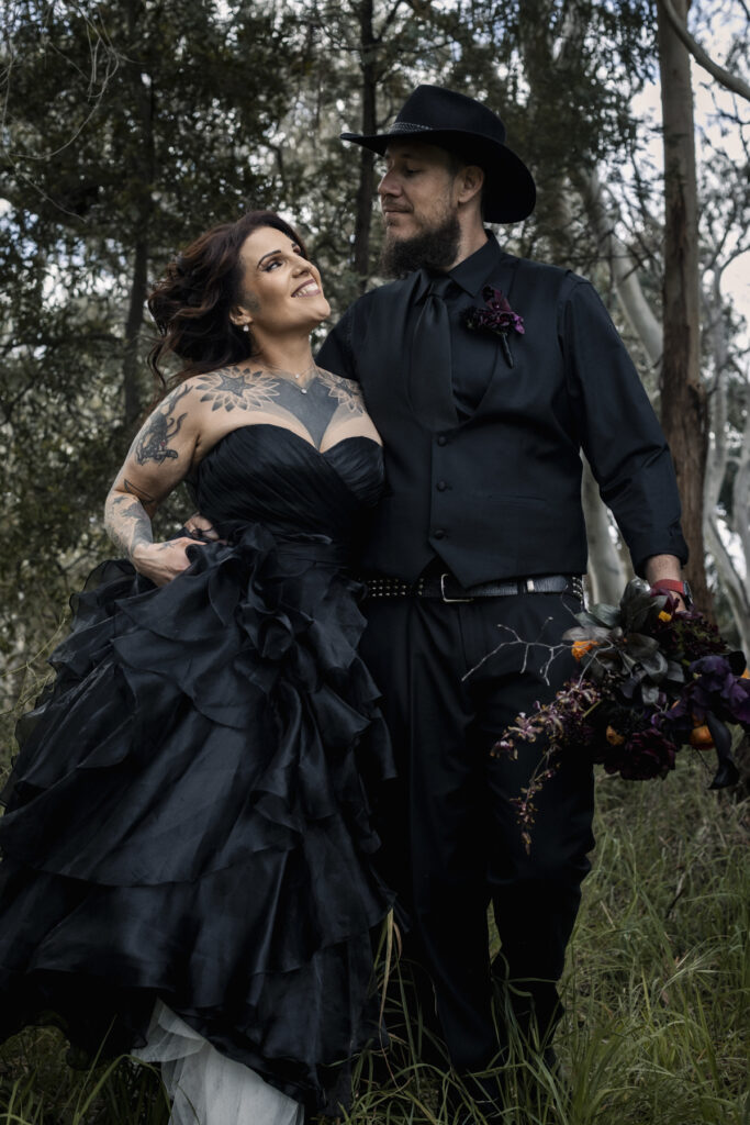 A bride and groom in black outfits, the bride is in a black ruffle wedding dress with dark moody flowers. The couple are more alternative with tattoos.