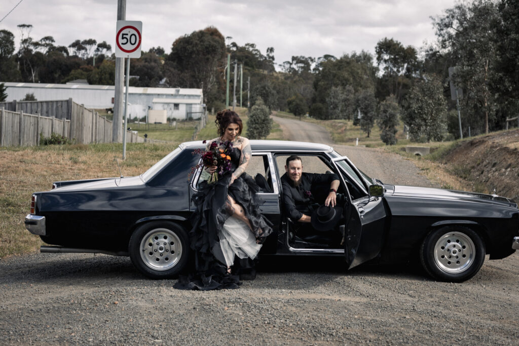 A bride and groom in black car, the bride is in a black ruffle wedding dress with dark moody flowers. The couple are more alternative with tattoos.