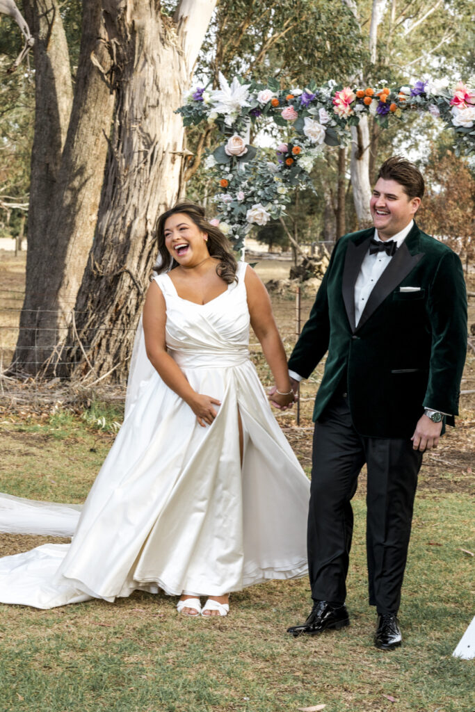 A couple laughing during their wedding ceremony at Paramoor Winery. The bride is in a beautiful white gown and the groom in in a stylish velvet green tuxedo jacket. They are standing under a floral arbour.