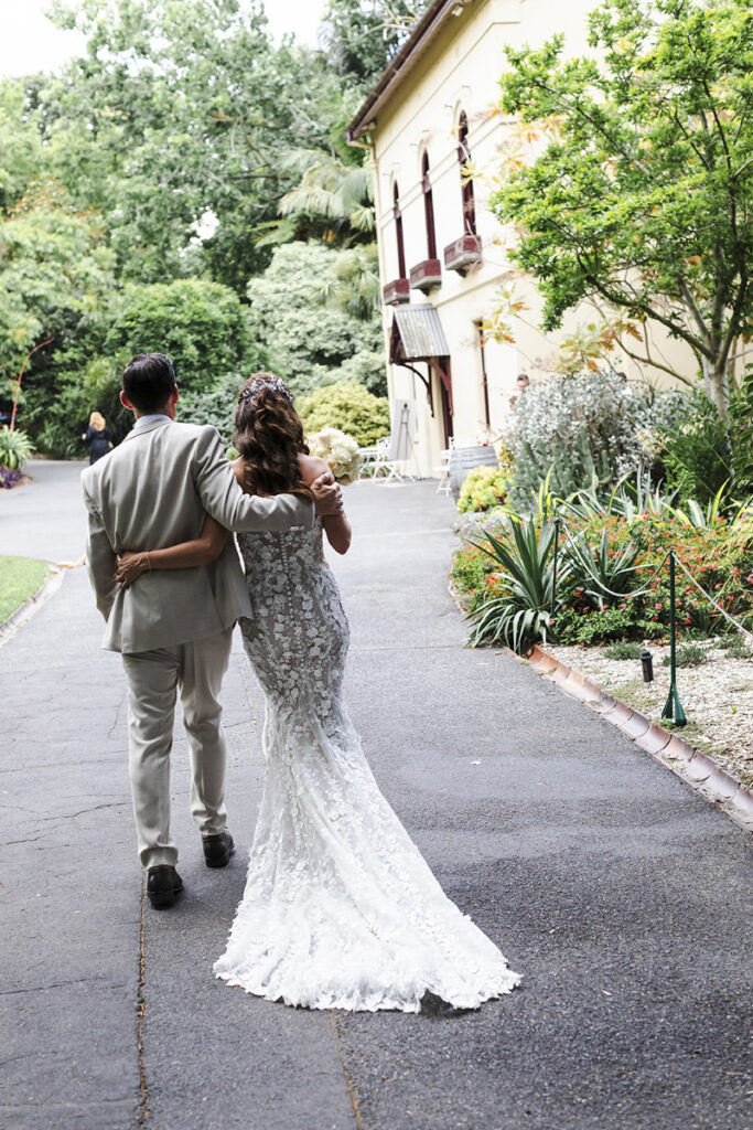 Gardens house wedding in Melbourne. A bride in a white dress with flowers with her groom in a tan suit, walking in front of gardens house in the Botanical gardens in Melbourne. They are looking at each other, it's a candid documentary style wedding image.