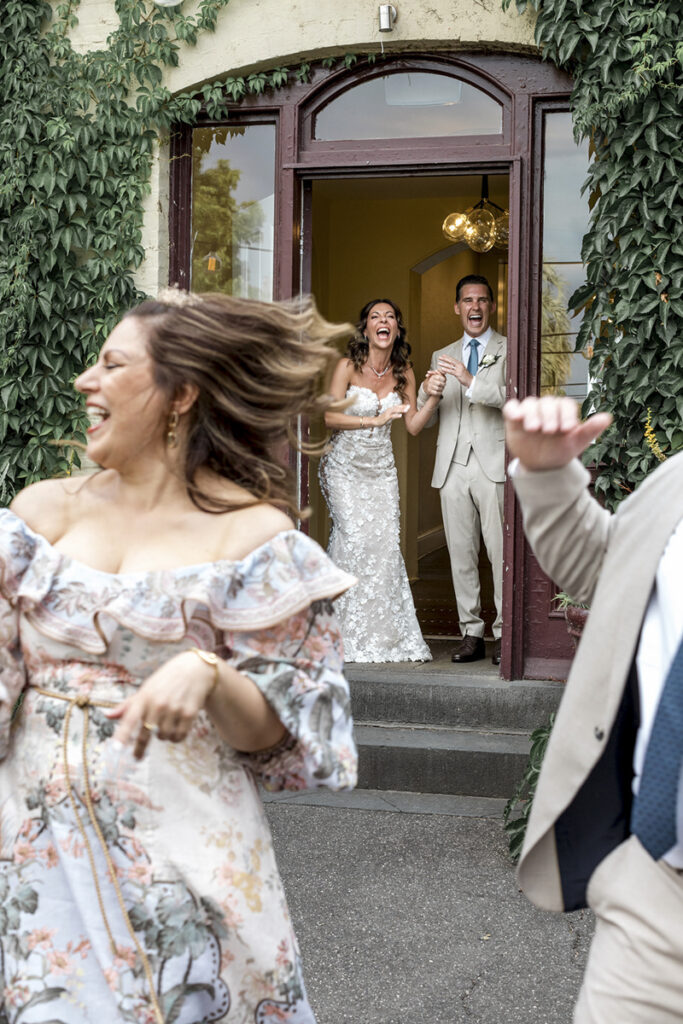 Gardens house wedding in Melbourne. A bride in a white dress with flowers with her groom in a tan suit, walking in front of gardens house in the Botanical gardens in Melbourne. They are laughing at the bridesmaid and groomsman dancing.